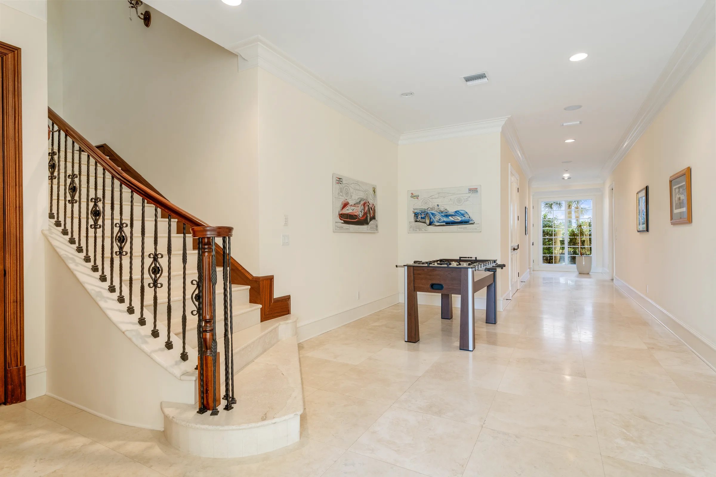 Foosball table in a long marble corridor with curved staircase, arched windows, and framed racing-car art