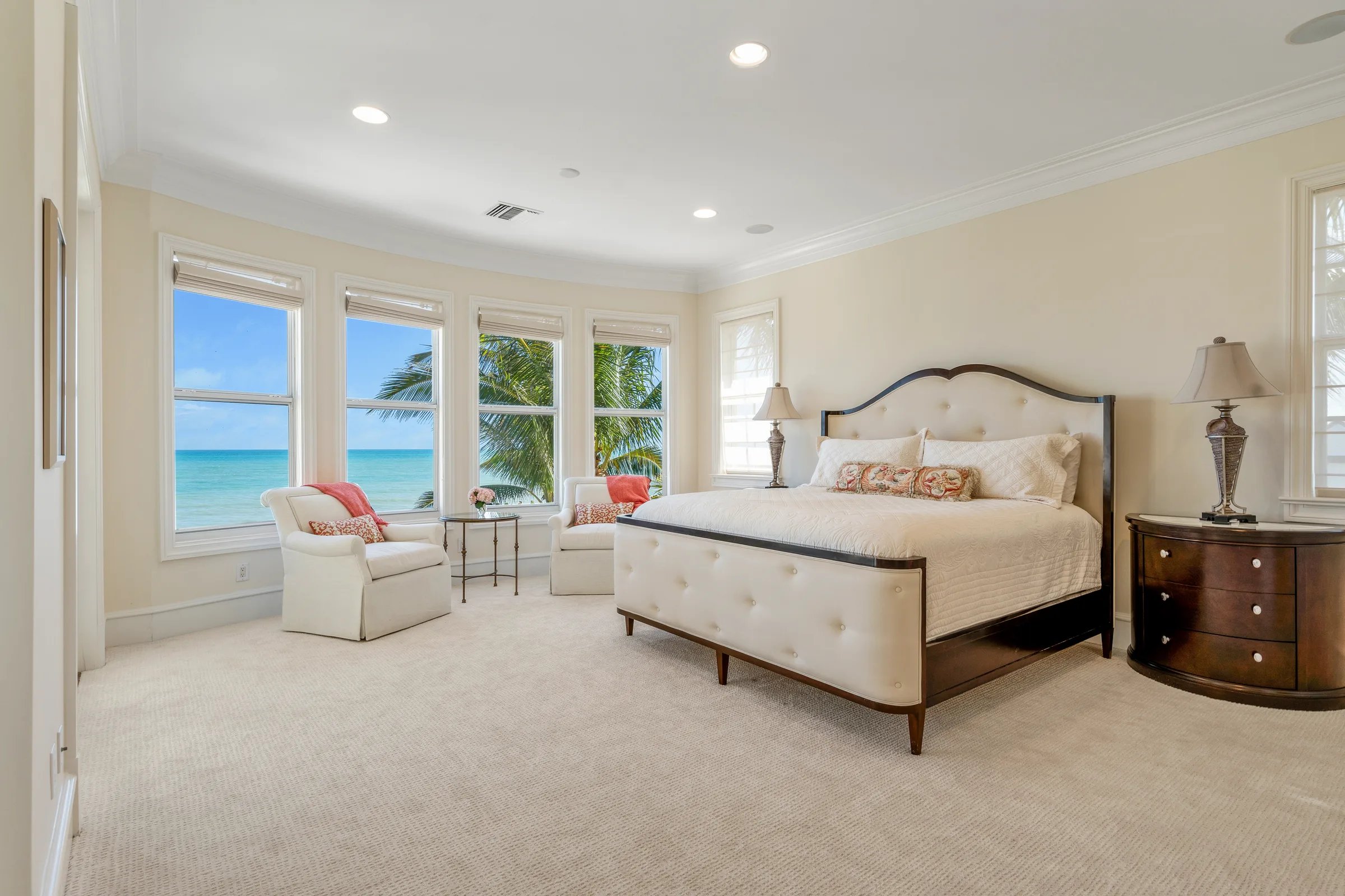 Guest bedroom with tufted king bed, mahogany nightstand, and a wall of bay windows opening to the Atlantic and palms