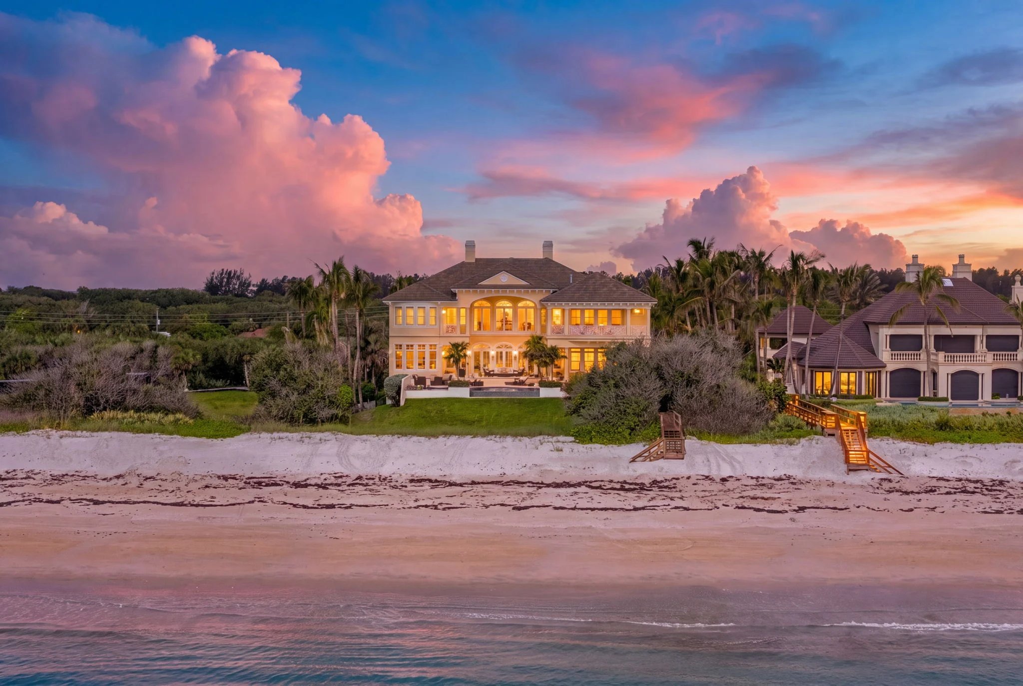 211 Beachside Drive at twilight from the Atlantic, the home rising from the dune line with lit windows, pink-cumulus sky, beach in the foreground